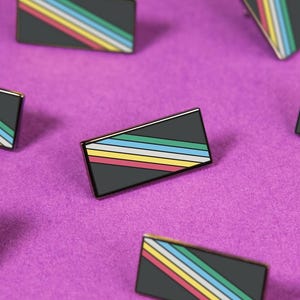 May include: A collection of rectangular enamel pins. Each pin has a black base with a diagonal stripe design in rainbow colours. The pins are scattered on a purple background.