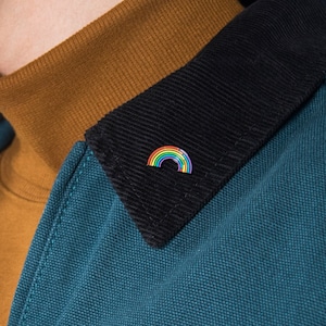 May include: A person wearing a teal blue jacket with a black corduroy collar. The jacket has a zipper closure and a rainbow pin on the collar.