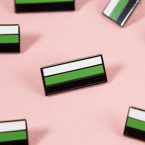 以下が含まれることがあります： 旗の形をした緑、白、黒のエナメルピンが複数あります。ピンはピンクの背景に散らばっています。