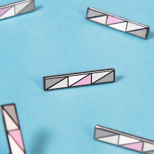 Puede incluir: Un conjunto de diez pines de esmalte con un diseño geométrico con triángulos rosas y grises. Los pines están dispuestos sobre un fondo azul claro.