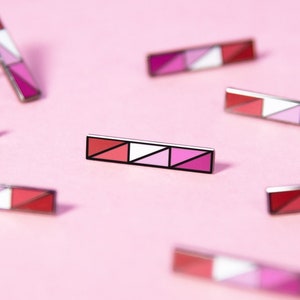 May include: Multiple pink, white, and red enamel pins with a geometric design. The pins are scattered on a pink background.