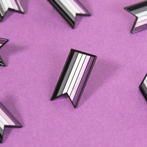 Op de afbeelding: Meerdere zwarte en witte emaille pinnen met een ontwerp van paarse, witte en zwarte strepen. De pinnen zijn gevormd als vlaggen en zijn gerangschikt op een paarse achtergrond.