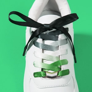May include: White sneaker with gradient green and gray shoelaces and a black bow. The laces are secured with a green lace lock. The text "MATCHING LACE LOCK + SHOELACE SET" is at the bottom.