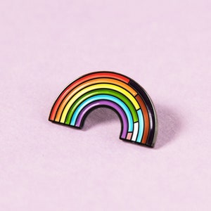 Può includere: Una spilla smaltata a forma di arcobaleno con un contorno nero. L'arcobaleno presenta bande di rosso, arancione, giallo, verde, blu, viola e una sezione con i colori della bandiera del Pride Progressista. La spilla è su uno sfondo rosa chiaro.