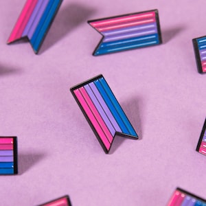 May include: A collection of 10 small, enamel pins in the shape of a bi-flag. The pins are arranged in a scattered pattern on a light purple background.