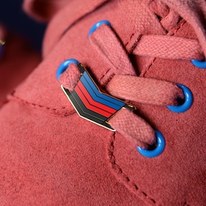 Puede incluir: Primer plano de un par de zapatos de gamuza rosa con cordones azules y un pin de colores arcoíris en los cordones.