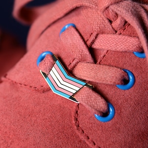 Könnte beinhalten: Ein Paar rote Wildlederschuhe mit blauen Schnürsenkeln und einer Transgender-Pride-Flaggennadel an einem Schuh.