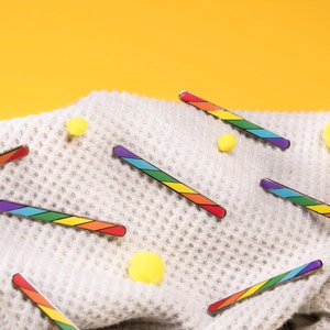 May include: A collection of rainbow-striped hair clips scattered on a textured, light gray blanket. Yellow pom-poms are also scattered on the blanket. The background is a solid yellow.