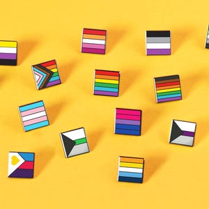 May include: A collection of square enamel pins featuring various LGBTQ+ pride flags. The pins are arranged on a bright yellow background. Each pin displays a unique flag design with vibrant colors, including rainbow, pink, blue, and black.