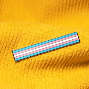 May include: A transgender pride pin with blue, pink, and light blue stripes on a yellow corduroy background.