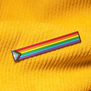 Könnte beinhalten: Eine regenbogenfarbene Emaille-Anstecknadel in Form eines Dreiecks. Die Anstecknadel befindet sich auf einem gelben Cordstoff.