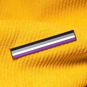 May include: A rectangular pin featuring the asexual pride flag, with horizontal stripes in black, gray, white, and purple. The pin is set against a textured, golden-yellow fabric background.