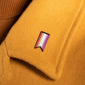 May include: A mustard yellow coat with a brown turtleneck. A small enamel pin with a flag design in shades of purple, white, red, and black is pinned to the coat's lapel. The coat's texture is soft and the overall aesthetic is warm and inviting.