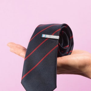 Pode incluir: Uma gravata azul escura com listras vermelhas está enrolada e sendo segurada em uma mão. Um prendedor de gravata prateado está preso à gravata.