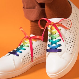 May include: White sneakers with rainbow-colored laces. The shoes have a perforated design and are set against an orange background. The laces are tied in bows, adding a pop of color to the footwear.