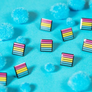 May include: A collection of small, colorful pins with a horizontal stripe pattern on a blue background. The pins are scattered amongst blue pom-poms.