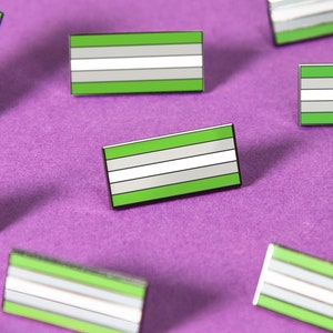 May include: Nine green, white, and gray enamel pins in the shape of a flag. The pins are scattered on a purple surface.