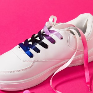 May include: White sneaker with gradient blue, purple, and pink ribbon laces. The laces transition from blue to purple and pink. The shoe is on a bright pink background with small pink pom-poms scattered around it.