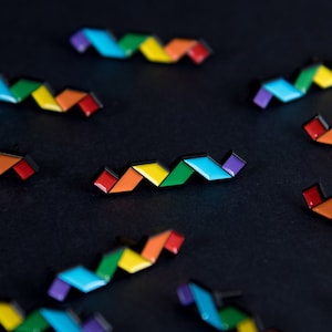 May include: A collection of rainbow enamel pins in the shape of a zig-zag pattern. The pins are scattered on a black surface.