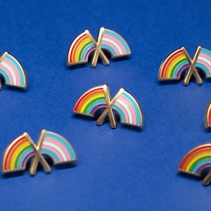 May include: A collection of enamel pins featuring a rainbow and transgender flag design. The pins have a gold outline and are set against a vibrant blue background. The rainbow flag displays the colors of the LGBTQ+ community.