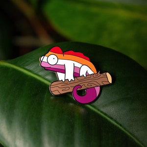 May include: A colorful enamel pin of a cartoon chameleon, perched on a brown log. The chameleon features the colors of the lesbian pride flag: orange, red, white, and purple. The pin is set against a backdrop of large, green leaves.