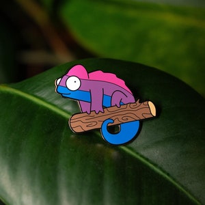 May include: A blue and pink enamel pin of a chameleon perched on a brown branch. The pin is a whimsical and colourful representation of a chameleon.