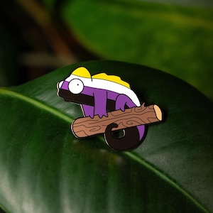 Może przedstawiać: Emaliowana przypinka przedstawiająca kreskówkowego kameleona w kolorze fioletowym, żółtym i białym, siedzącego na gałęzi. Kameleon ma duże, okrągłe oko.