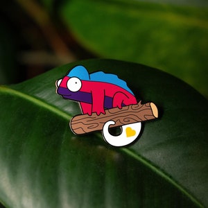 Può includere: Un personaggio di cartone animato di camaleon blu, rosso e viola seduto su un tronco marrone con un cuore giallo. Il personaggio è uno spillo su una foglia verde.