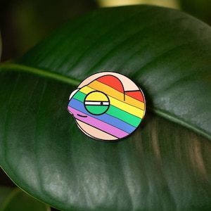 Może przedstawiać: Emaliowana szpilka w kształcie tęczy z uśmiechniętą buzią i tęczowym sercem. Szpilka znajduje się na zielonym liściu.