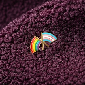 May include: A gold-colored pin featuring two crossed flags. One flag is a rainbow, and the other is the transgender pride flag. The pin is set against a textured, deep purple fabric background.