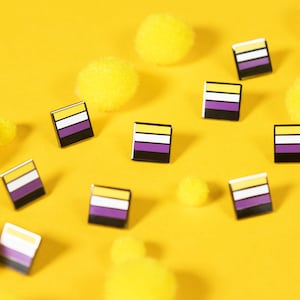 Op de afbeelding: Een gele achtergrond met meerdere gele pompoenen en 10 kleine vierkante pinnen met de kleuren van de non-binaire pride-vlag: geel, wit, paars en zwart.