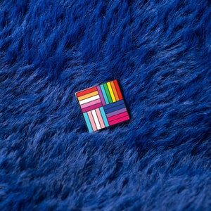 Może przedstawiać: Kwadratowa emaliowana przypinka z kolorowym geometrycznym wzorem, przedstawiającym kolory flagi dumy LGBTQ+. Przypinka jest umieszczona na tle głębokiej niebieskiej, puszystej tkaniny.