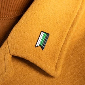 Peut inclure: Gros plan sur une veste moutarde avec une petite épingle émaillée. L'épingle présente un motif de drapeau à rayures vertes et blanches. La veste est portée sur un pull à col roulé marron.