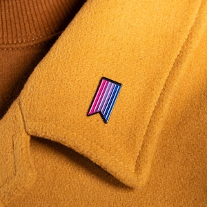 May include: A gold-colored wool coat with a brown turtleneck. A small enamel pin with the bisexual pride flag colors of pink, purple, and blue is pinned to the coat's lapel. The pin is a small rectangle with a black border.