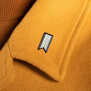 Peut inclure: Une petite épingle en émail avec un bord noir et un motif de rayures blanches, vertes et jaunes. L'épingle est fixée au revers d'un manteau jaune moutarde. Un pull à col roulé marron est visible en dessous.
