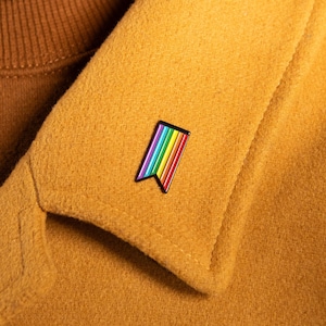 May include: A rainbow flag pin is attached to a mustard-colored coat. The pin features a colorful design with red, orange, yellow, green, blue, and purple stripes. The coat is worn over a brown turtleneck.
