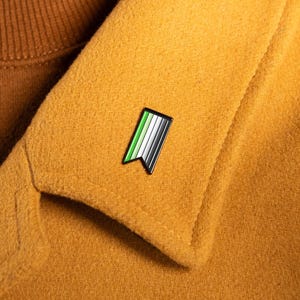 May include: A close-up of a mustard-colored coat with a small pin attached to the lapel. The pin features a design with green, white, black, and gray stripes. The person is wearing a brown turtleneck sweater.