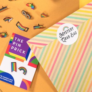 Può includere: Un assortimento di spille a tema arcobaleno, un nastro a righe e una busta "Mystery Grab Bag" su una superficie gialla. Le spille presentano vari disegni, tra cui arcobaleni, bandiere e forme geometriche. Una carta recita "The Pin Prick".