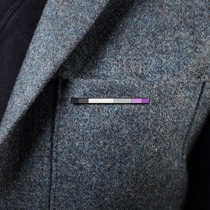 May include: A grey blazer with a small pin featuring a black, white, grey, and purple striped design.