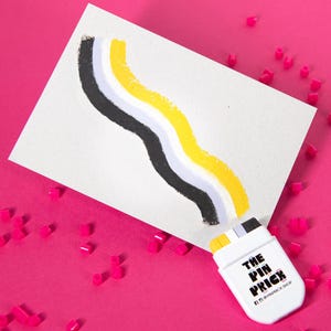 May include: A white card with a black, gray, and yellow wavy design, next to a white marker with the text "THE PIN PRICK". The background is a bright pink with small pink cube-shaped objects scattered around.