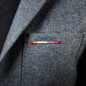 Puede incluir: Una chaqueta gris con un pequeño pin en el bolsillo. El pin tiene un diseño de rayas de arcoíris.