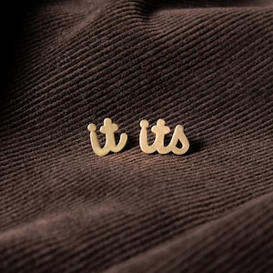 May include: A pair of gold earrings with the word "it's" written on them. The earrings are on a brown fabric background.