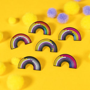 May include: Seven rainbow enamel pins on a yellow background. The pins are in the shape of an arc and feature different coloured stripes. The pins are surrounded by small yellow and blue pom-poms.