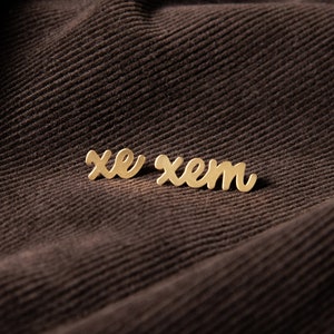 May include: Gold-colored metal letters spelling out the word "xem" on a brown corduroy fabric background.