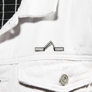 May include: A silver pin shaped like an archway with a textured design. The pin is on a white denim jacket.
