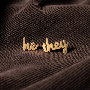 May include: Gold-colored metal earrings with the words "he they" written in cursive.