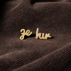 May include: Gold-colored metal earrings with the words "je hur" written in cursive.