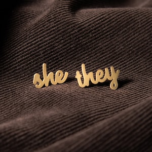 Pode incluir: Dois brincos de metal dourado com as palavras "she" e "they" escritas em letra cursiva. Os brincos estão sobre um fundo de tecido marrom.