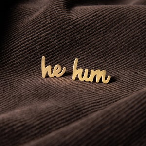 Op de afbeelding: Een goudkleurige speld met de woorden "he hum" in een cursief lettertype. De speld is geplaatst tegen een donkerbruine corduroy stoffen achtergrond. De speld is klein en delicaat, met een glanzende afwerking.