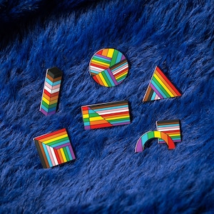 May include: A set of seven rainbow-coloured enamel pins in various geometric shapes, including a circle, a rectangle, a triangle, and a curved shape. The pins are arranged on a blue fuzzy surface.